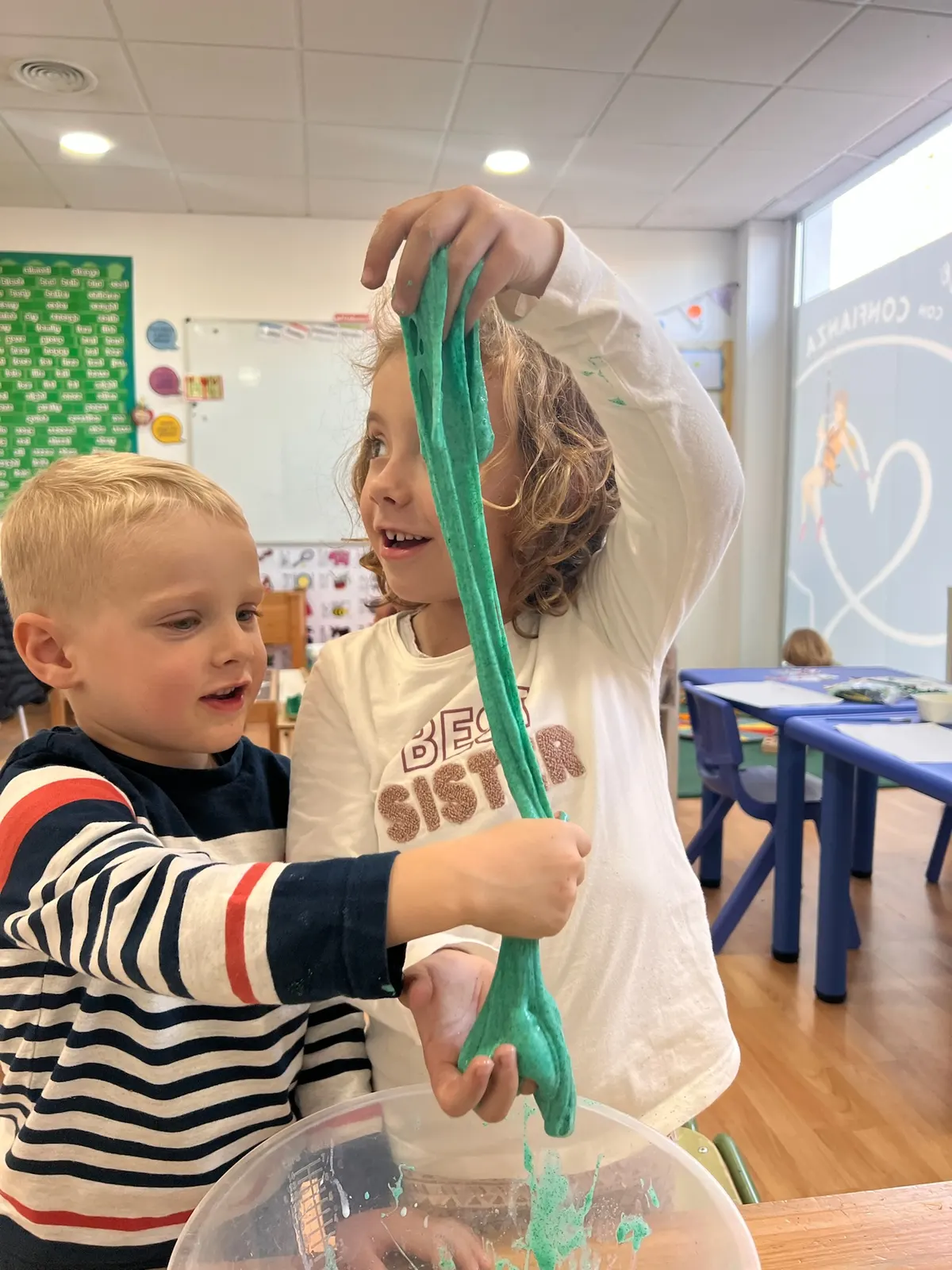 Dos niños riéndose mientras estiran slime verde brillante sobre un cuenco en el aula