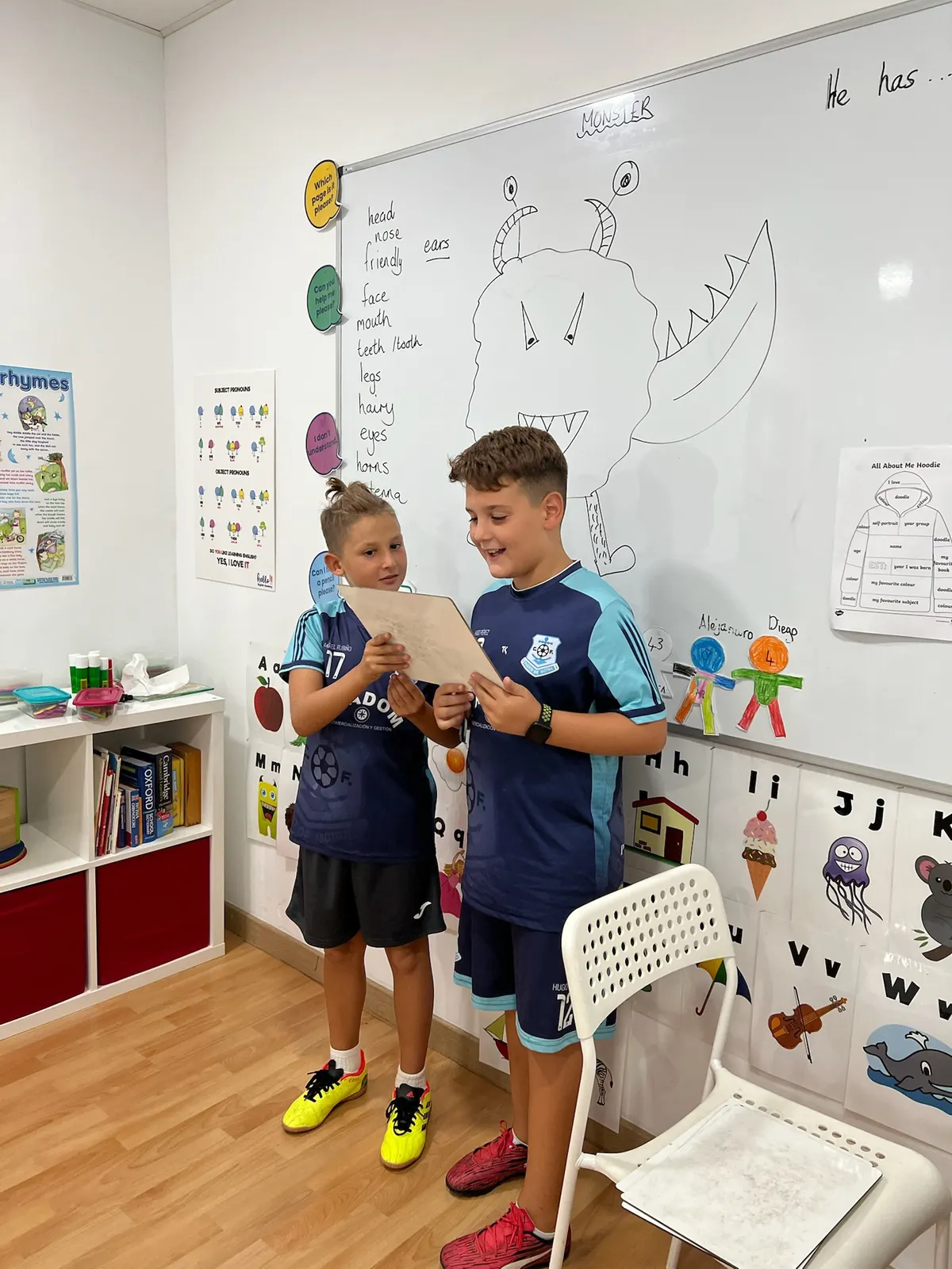 Dos niños con equipación de fútbol leyendo en voz alta la descripción de su monstruo ante una pizarra con vocabulario del cuerpo en inglés