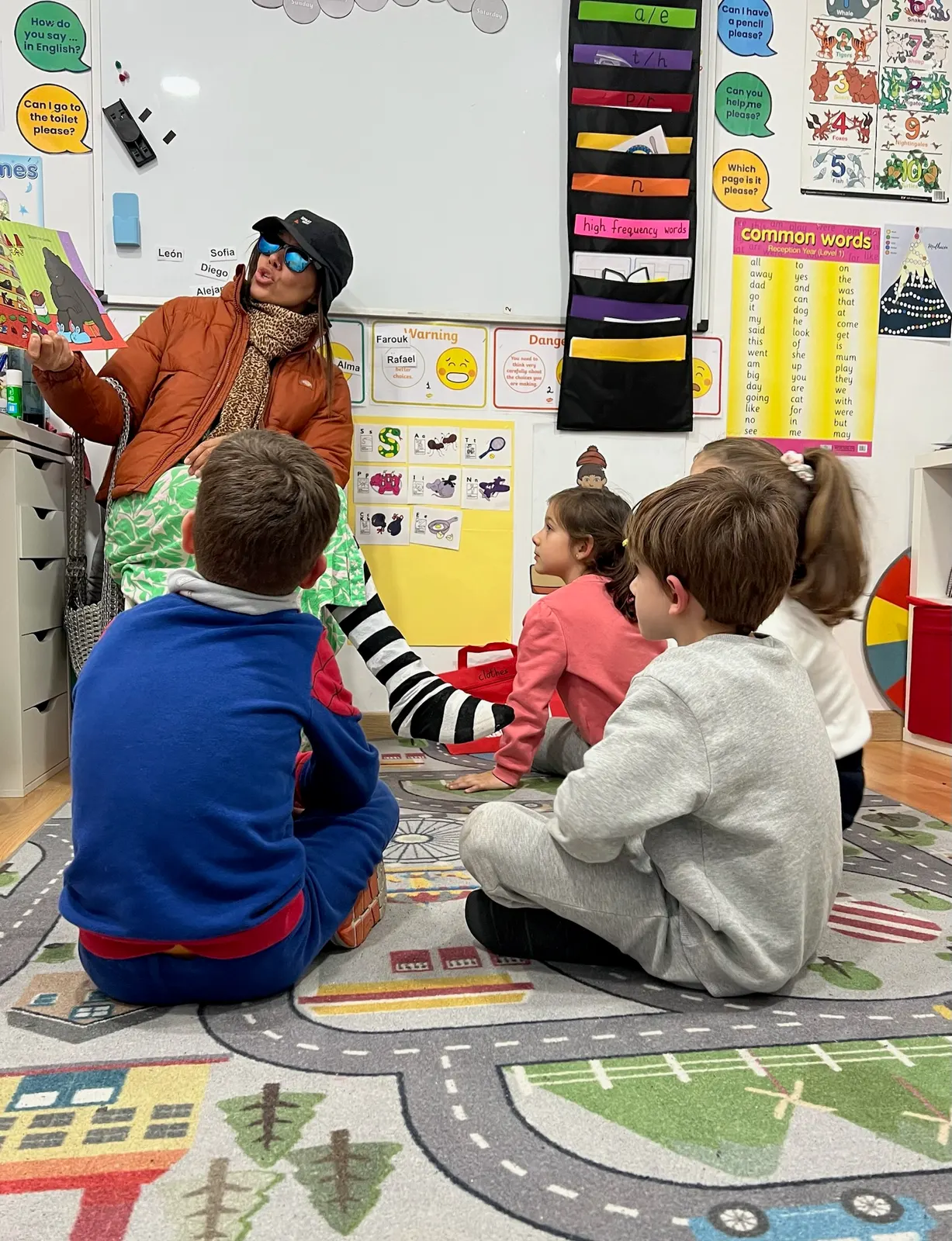 Profesora con chaqueta naranja de disfraz, gorra y gafas de sol leyendo un cuento a niños sentados sobre una alfombra de carreteras