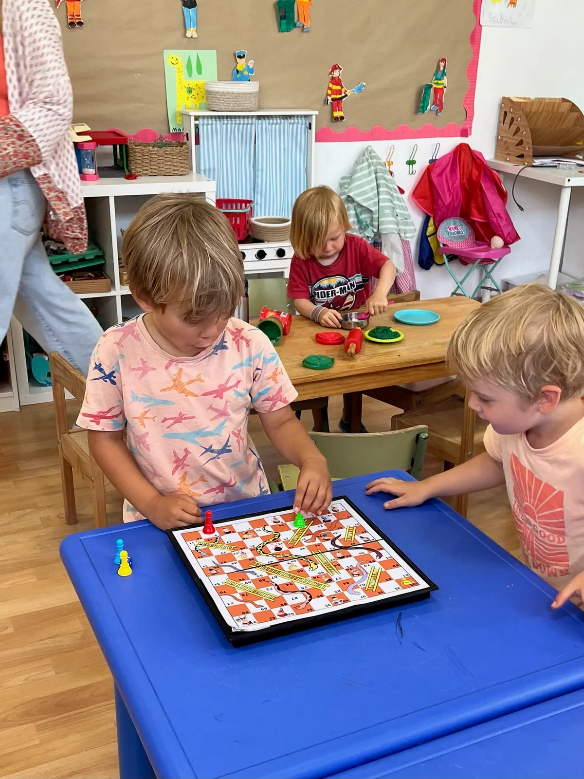 Three young children playing snakes and ladders together at a blue table in the classroom, painted paper figures on the wall behind