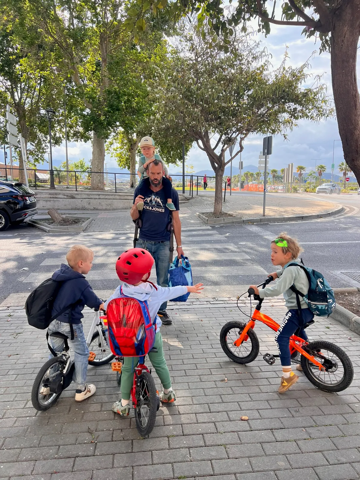Ella's English school-run scene: a parent carrying a toddler on his shoulders as three children arrive on small bikes at the academy