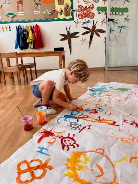 Young child painting a large collaborative mural on paper on the classroom floor, insect artwork on the wall behind