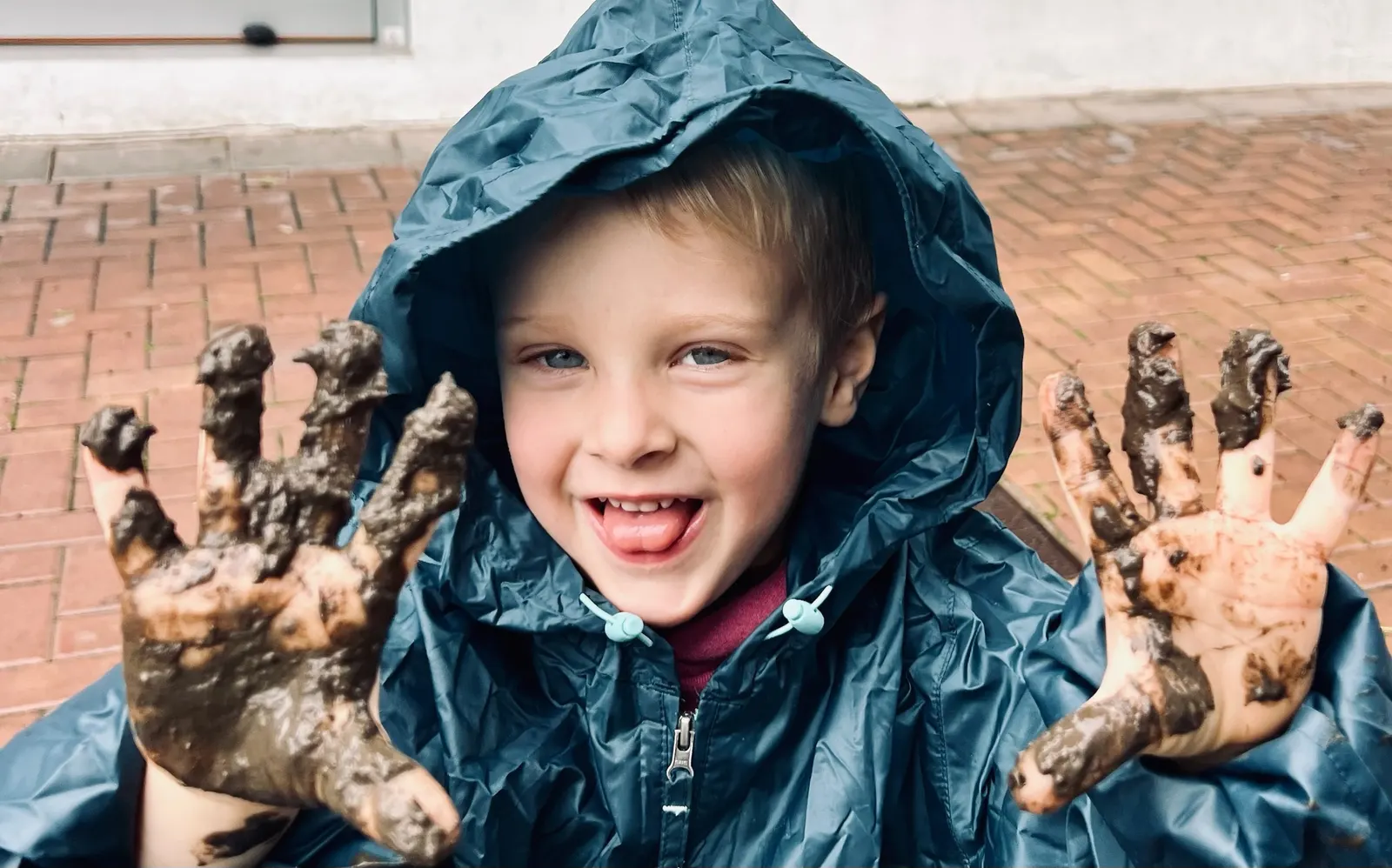 Boy in a navy raincoat showing two hands covered in mud after rainy-day play