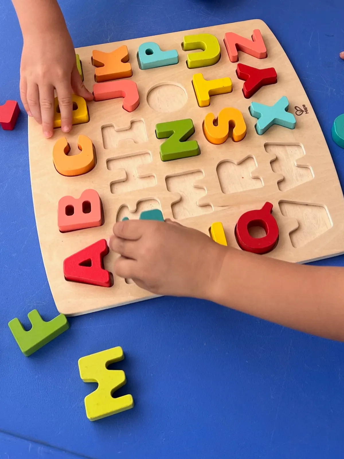 Manos de niños colocando letras de madera en un puzle del abecedario sobre una mesa azul