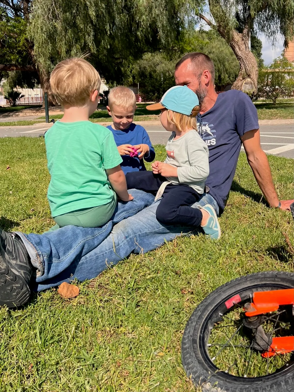 Profesor sentado en el césped de un parque con un grupo pequeño de tres niños y una bicicleta sin pedales naranja