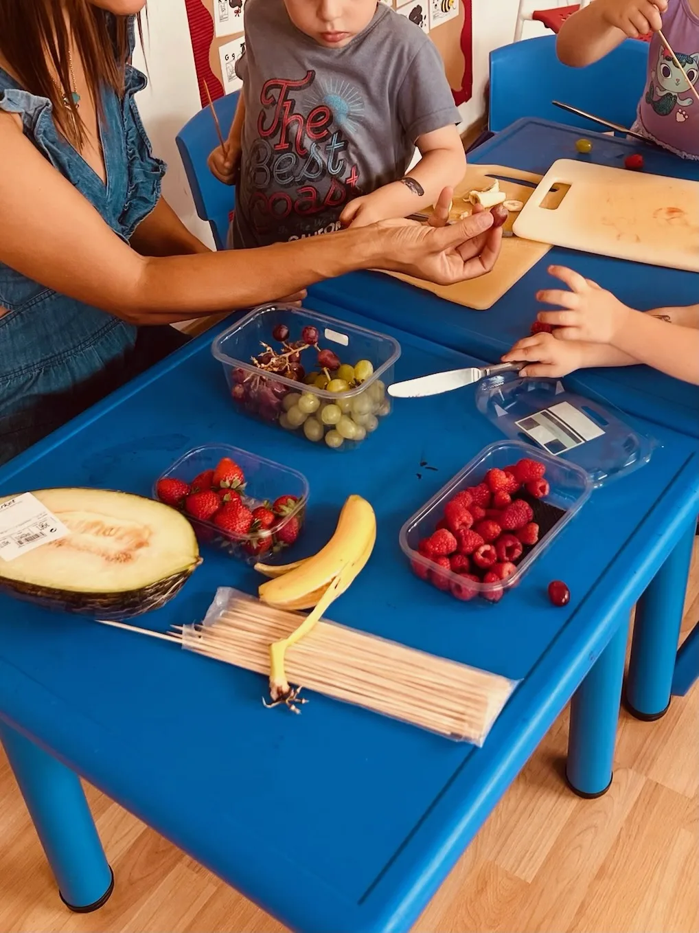 Children making fruit skewers with a teacher at the classroom table
